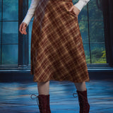 Woman wearing brown plaid A-line midi skirt with braided belt and hand in side pocket, styled with white blouse and boots