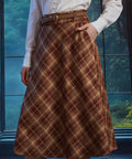 Woman wearing a brown plaid high-waisted midi skirt with braided belt, A-line silhouette and hand in side pocket