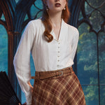 Woman wearing an ivory pearl button blouse with shawl collar and long sleeves, styled with a brown plaid skirt and braided belt