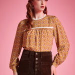 Woman wearing brown floral prairie blouse with lace trim and long sleeves against pink background