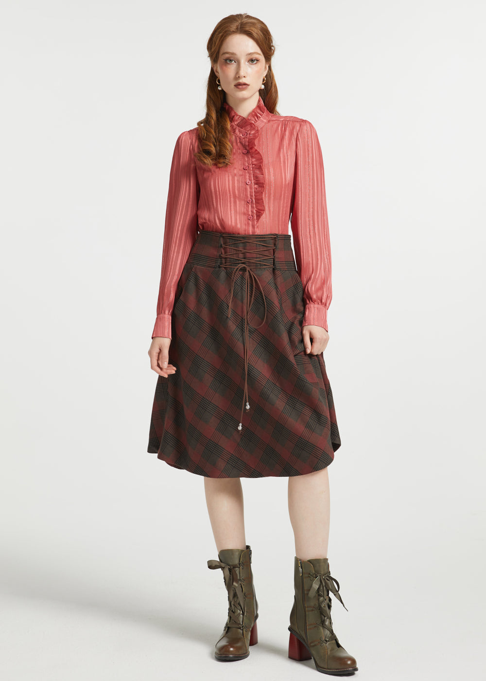 A model stands against a light gray studio background wearing a long-sleeve pink pleated blouse, a dark red and black plaid skirt with a drawstring waist, and olive green lace-up ankle boots.