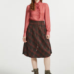 A model stands against a light gray studio background wearing a long-sleeve pink pleated blouse, a dark red and black plaid skirt with a drawstring waist, and olive green lace-up ankle boots.