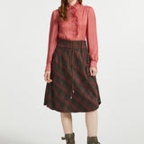 A model stands against a light gray studio background wearing a long-sleeve pink pleated blouse, a dark red and black plaid skirt with a drawstring waist, and olive green lace-up ankle boots.