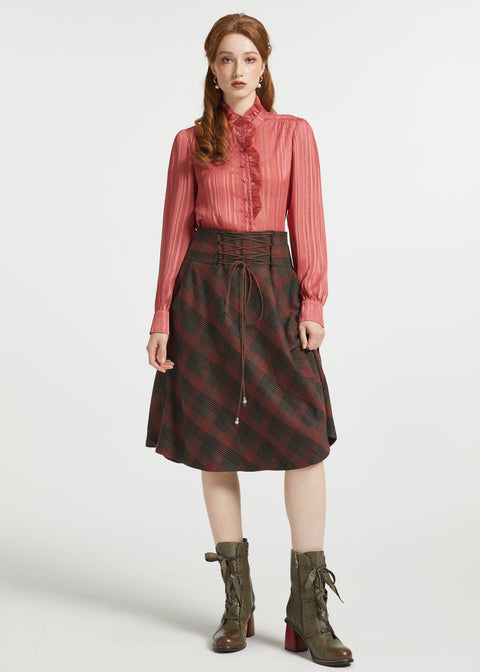 A model stands against a light gray studio background wearing a long-sleeve pink pleated blouse, a dark red and black plaid skirt with a drawstring waist, and olive green lace-up ankle boots.