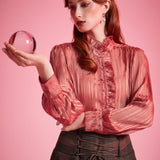 Woman wearing a dusty rose striped ruffle blouse with high neckline, button front, and long blouson sleeves while holding a glass sphere