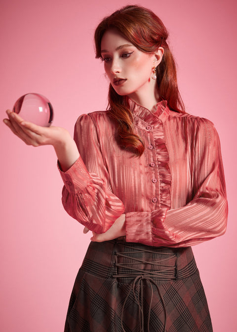 Woman wearing a dusty rose striped ruffle blouse with high neckline, button front, and long blouson sleeves while holding a glass sphere