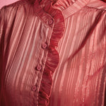 Close-up of dusty rose ruffle blouse showing the high ruffled neckline, button front, gathered shoulder, and shimmering tonal stripes