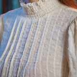 Close-up of cream color textured blouse showing smocked ruffle neckline, front pintucks and raised dobby weave