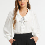 Front view of woman wearing ivory long sleeve blouse with polka dot necktie and black trousers