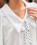 Close-up of ivory blouse with oversized pointed collar, pom-pom trim and black polka dot necktie