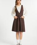 Full-length studio view of woman wearing a brown pleated jumper dress with ivory lace blouse, white socks and burgundy Mary Jane shoes.