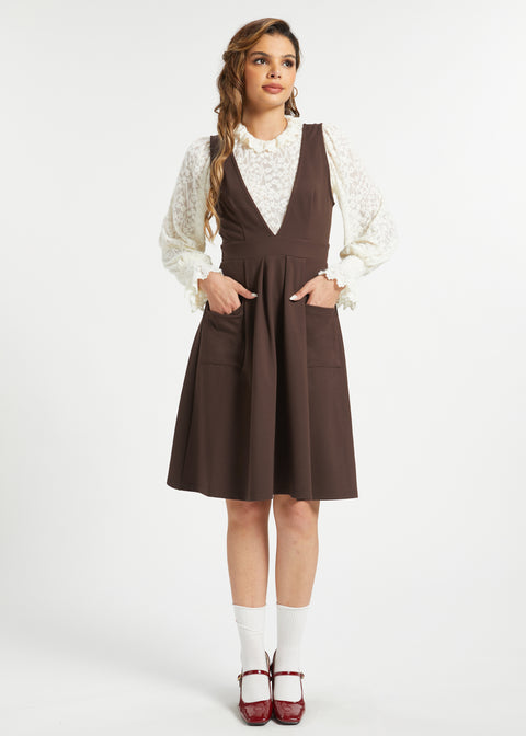 Full-length studio view of woman wearing a brown pleated jumper dress with ivory lace blouse, white socks and burgundy Mary Jane shoes.