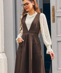 Woman modeling the brown V-neck pinafore jumper dress over an ivory lace blouse with one hand in the front patch pocket.