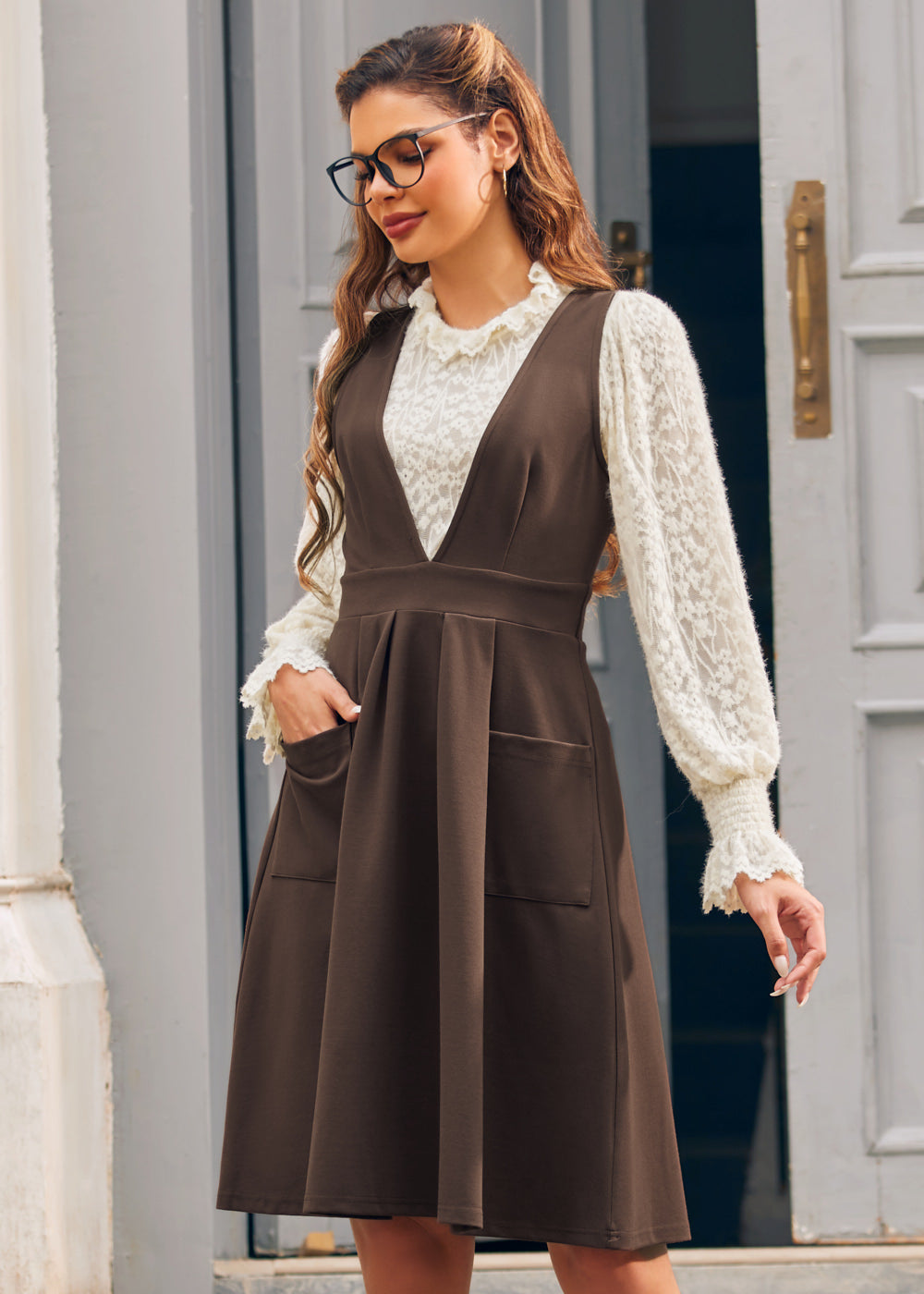Woman modeling the brown V-neck pinafore jumper dress over an ivory lace blouse with one hand in the front patch pocket.