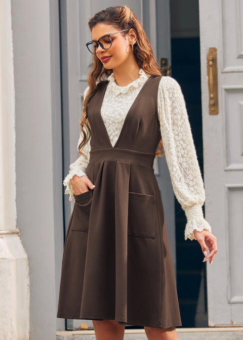 Woman modeling the brown V-neck pinafore jumper dress over an ivory lace blouse with one hand in the front patch pocket.