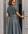 Back view of woman wearing blue floral cotton dress showing fitted shirt bodice, full skirt and sage green belt