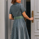 Back view of woman wearing blue floral cotton dress showing fitted shirt bodice, full skirt and sage green belt