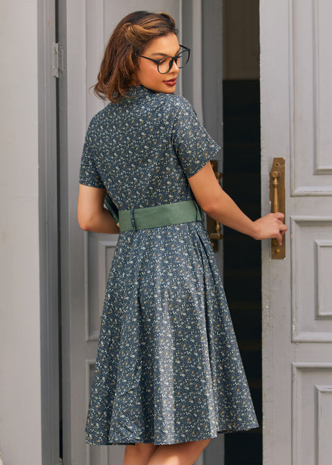 Back view of woman wearing blue floral cotton dress showing fitted shirt bodice, full skirt and sage green belt