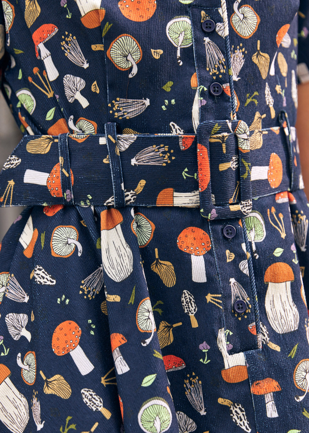 Close-up of a navy blue fabric shirtdress with an allover mushroom print in orange, white, beige, and green, featuring a matching fabric belt with a covered buckle.