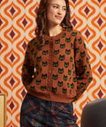 A woman wearing a brown button-front cardigan with a repeating black cat face pattern, paired with dark plaid pants, posed against a retro orange and cream patterned wall.