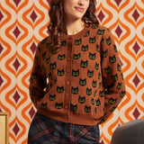 A woman wearing a brown button-front cardigan with a repeating black cat face pattern, paired with dark plaid pants, posed against a retro orange and cream patterned wall.