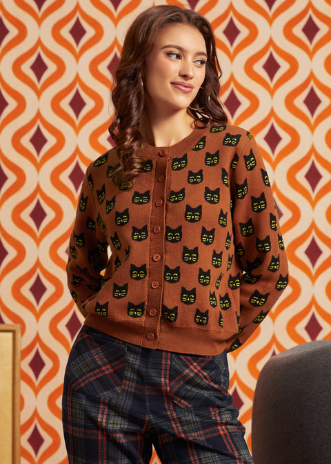 A woman wearing a brown button-front cardigan with a repeating black cat face pattern, paired with dark plaid pants, posed against a retro orange and cream patterned wall.