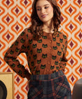 A woman wearing a brown button-front cardigan with a black cat face pattern, paired with dark plaid trousers, posed against a retro orange and cream patterned wall.