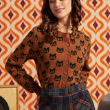 A woman wearing a brown button-front cardigan with a black cat face pattern, paired with dark plaid trousers, posed against a retro orange and cream patterned wall.