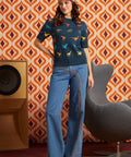 A woman stands in a retro-styled room wearing a navy blue short-sleeve knit top with colorful dinosaur prints, and blue wide-leg jeans.