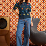 A woman stands in a retro-styled room wearing a navy blue short-sleeve knit top with colorful dinosaur prints, and blue wide-leg jeans.