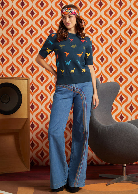 A woman stands in a retro-styled room wearing a navy blue short-sleeve knit top with colorful dinosaur prints, and blue wide-leg jeans.