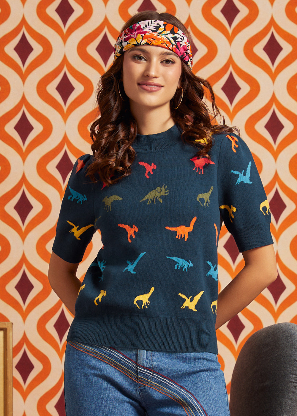 A woman wearing a navy blue short-sleeve knit sweater with a colorful dinosaur print, paired with blue jeans and a patterned headband, standing in front of a retro orange and cream patterned wall.