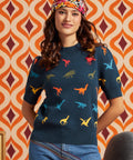 A woman wearing a navy blue short-sleeve knit sweater with a colorful dinosaur print, paired with blue jeans and a patterned headband, standing in front of a retro orange and cream patterned wall.