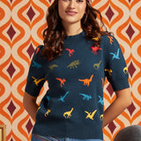 A woman wearing a navy blue short-sleeve knit sweater with a colorful dinosaur print, paired with blue jeans and a patterned headband, standing in front of a retro orange and cream patterned wall.