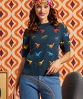A woman wearing a navy blue short-sleeve knit sweater with a colorful dinosaur print, paired with blue jeans and a patterned headband, standing in front of a retro orange and cream patterned wallpaper background.