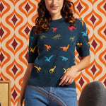 A woman wearing a navy blue short-sleeve knit sweater with a colorful dinosaur print, paired with blue jeans and a patterned headband, standing in front of a retro orange and cream patterned wallpaper background.