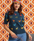 A woman wearing a navy blue sweater with a colorful dinosaur print, styled with a floral headband and blue jeans, set against a retro orange and brown patterned wall.