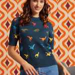 A woman wearing a navy blue sweater with a colorful dinosaur print, styled with a floral headband and blue jeans, set against a retro orange and brown patterned wall.