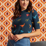 A woman wearing a navy blue short-sleeve top with a colorful dinosaur print, styled with a floral headband and blue jeans, set against a retro orange and brown patterned background.