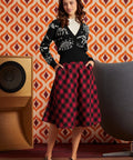A woman stands in a retro-styled room wearing a black cardigan with white skeleton graphics and a red-and-black buffalo plaid midi skirt. She is posed in front of a bold orange, brown, and white geometric wallpaper, with a wooden speaker on the left and a gray chair on the right.