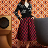 A woman stands in a retro-styled room wearing a black cardigan with white skeleton graphics and a red-and-black buffalo plaid midi skirt. She is posed in front of a bold orange, brown, and white geometric wallpaper, with a wooden speaker on the left and a gray chair on the right.