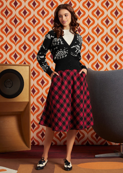 A woman stands in a retro-styled room wearing a black cardigan with white skeleton graphics and a red-and-black buffalo plaid midi skirt. She is posed in front of a bold orange, brown, and white geometric wallpaper, with a wooden speaker on the left and a gray chair on the right.
