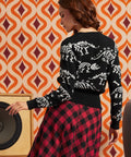 A woman shown from the back wearing a black knit sweater with white dinosaur skeleton patterns and a red-and-black plaid skirt, standing in front of a retro orange and cream patterned wall.