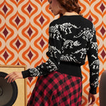 A woman shown from the back wearing a black knit sweater with white dinosaur skeleton patterns and a red-and-black plaid skirt, standing in front of a retro orange and cream patterned wall.