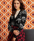 A woman wearing a black cardigan with white skeleton motifs over a cream ribbed top and a red-and-black plaid skirt, posed in front of an orange and cream geometric patterned wall.