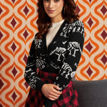 A woman wearing a black cardigan with white skeleton motifs over a cream ribbed top and a red-and-black plaid skirt, posed in front of an orange and cream geometric patterned wall.