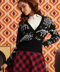 A woman wearing a black knit cardigan with white skeleton motifs, a white ribbed turtleneck, and a red and black checkered skirt, posed against a retro orange and cream patterned wallpaper background.