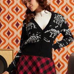 A woman wearing a black knit cardigan with white skeleton motifs, a white ribbed turtleneck, and a red and black checkered skirt, posed against a retro orange and cream patterned wallpaper background.