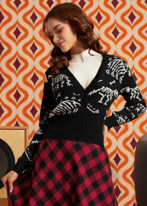 A woman wearing a black knit cardigan with white skeleton motifs, a white ribbed turtleneck, and a red and black checkered skirt, posed against a retro orange and cream patterned wallpaper background.