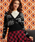 A woman wearing a black cardigan with white skeleton motifs over a white ribbed top and a red and black plaid skirt, posed against a retro orange and brown patterned wallpaper background.